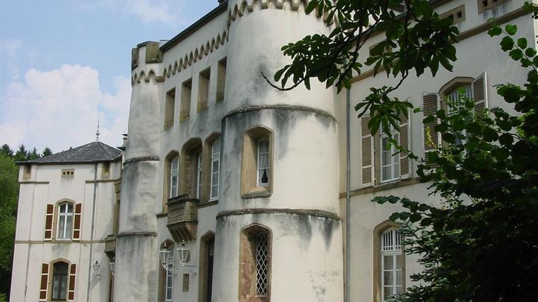 Ein historisches Gebäude mit einem turmähnlichen Anbau und dekorativen Fenstern. Grüne Bäume umgeben die elegante Architektur.