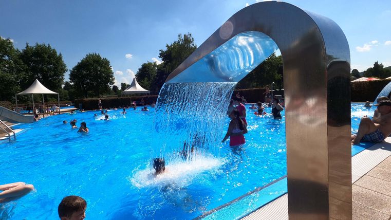 Une piscine avec de l'eau claire et bleue et de nombreux baigneurs. Une douche en cascade offre un rafraîchissement supplémentaire.