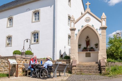 Zwei Männer sitzen auf einer Bank vor einem weißen Gebäude mit einer religiösen Statue in einer Nische.