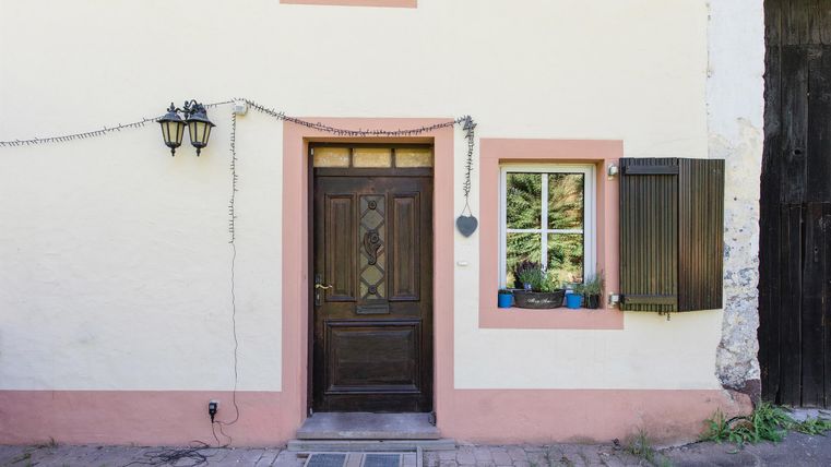 Ein gemütliches Haus mit einer holzfarbenen Tür und einem Fenster. Die Fassade ist in sanften Farben gestrichen und hat Blumen im Fenster.