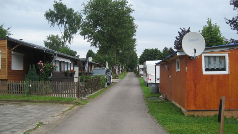 Een rustige straat met vakantiewoningen en groene weiden. Boven de hutten zijn enkele bomen en een bewolkte lucht te zien.
