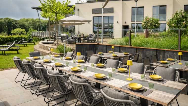 Un espace extérieur élégant avec une grande table dressée pour un repas. À l'arrière-plan, on aperçoit un bâtiment moderne et un jardin bien entretenu.