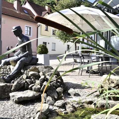 Bronzestatue eines Mannes auf Steinen, umgeben von Schilf und einem kleinen Teich. Im Hintergrund sind H&auml;user und ein Sonnenschirm zu sehen., &copy; Tourist-Information Bitburger Land
