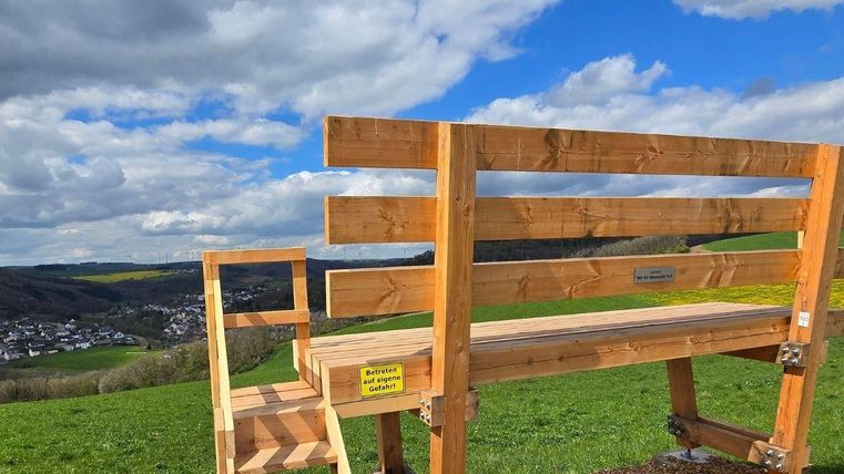 Een houten bank met trap staat op een heuvel met uitzicht op een landschap. De lucht is gedeeltelijk bewolkt en het groen van de weiden is goed zichtbaar.