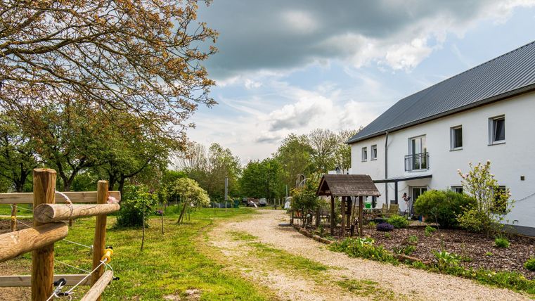 Eine ländliche Szenerie mit einem schmalen Weg, der zu einem modernen Haus führt. Im Hintergrund sind grüne Bäume und ein kleiner Spielplatz zu sehen.