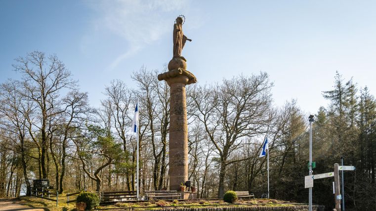 Eine Statue auf einer Säule steht in einer ruhigen Umgebung mit Bäumen. Vor der Statue sind Sitzbänke und Fahnen zu sehen.