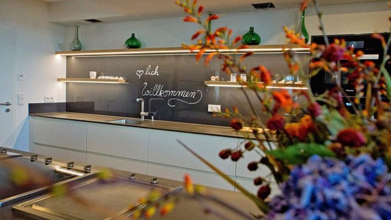 Une cuisine moderne avec des étagères élégantes et un tableau qui écrit "Bienvenue". Au premier plan, des arrangements floraux colorés sont visibles.