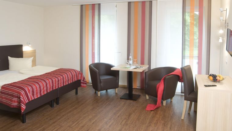 Une chambre moderne avec un lit double et des fauteuils confortables. De grandes fenêtres avec des rideaux colorés laissent entrer beaucoup de lumière.