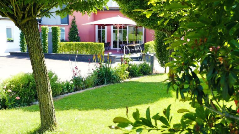 Ein schöner Garten mit gepflegtem Rasen und Bäumen. Im Hintergrund ist ein Haus mit einer Terrasse und Sonnenschirm zu sehen.