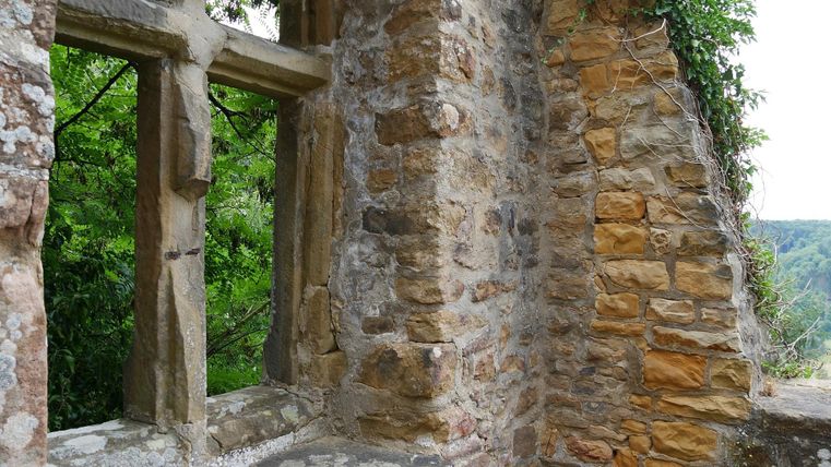 Eine Ruine mit einem großen Fenster aus Stein. Der Blick zeigt grüne Bäume und eine hügelige Landschaft im Hintergrund.