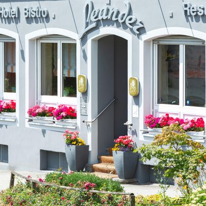 Un hôtel accueillant avec des fleurs colorées devant l'entrée. La façade est gris clair avec de grandes fenêtres.