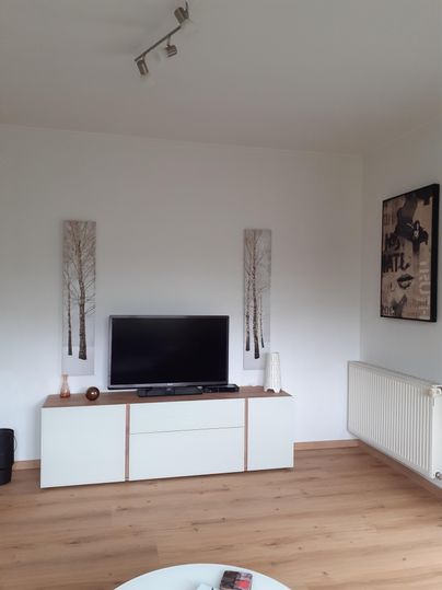 Un espace de vie moderne avec une télévision sur un meuble TV blanc. Aux murs, des images d'arbres et une autre œuvre d'art sont accrochées.