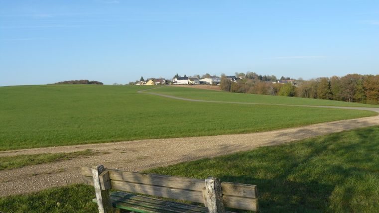 Vue d'une prairie verte avec un chemin et un banc au premier plan, à l'arrière-plan un village sous un ciel bleu.
