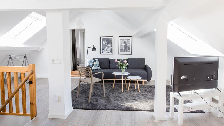 Ein heller und moderner Wohnraum mit einer gemütlichen Couch, einem Couchtisch und einem Flachbildfernseher. Große Fenster sorgen für viel Tageslicht und eine einladende Atmosphäre.