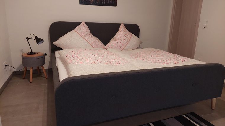 A modern bedroom with a large, cozy bed and decorative pillows. Next to the bed is a small table with a lamp and a rug in the background.