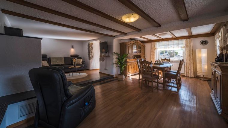 Ein gemütliches Wohnzimmer mit Holzdeckenbalken und einem Esstisch. Der Raum ist hell und einladend, mit einer großen Fensterfront und einem bequemen Sessel.