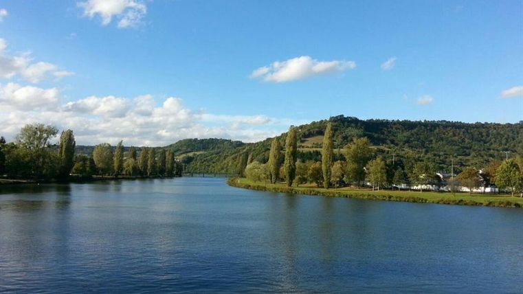 Ein ruhiger Fluss mit sanften Wellen und grünen Ufern. Im Hintergrund sind Hügel und ein blauer Himmel mit wenigen Wolken zu sehen.