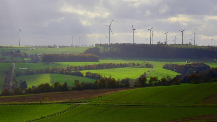 Een groene landschap met zachte heuvels en windturbines op de achtergrond. De lucht is bewolkt en er zijn zachte lichtstralen.
