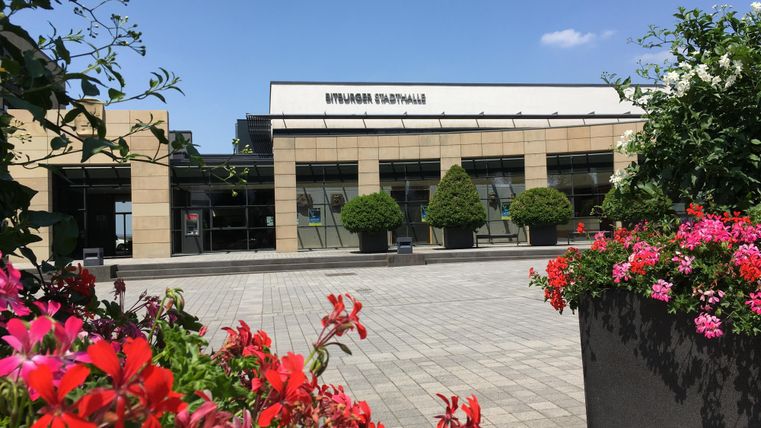 Ein modernes Gebäude mit großen Fenstern und gepflegten Pflanzen im Vordergrund. Bunte Blumenbeete verleihen der Umgebung Farbe und Leben.