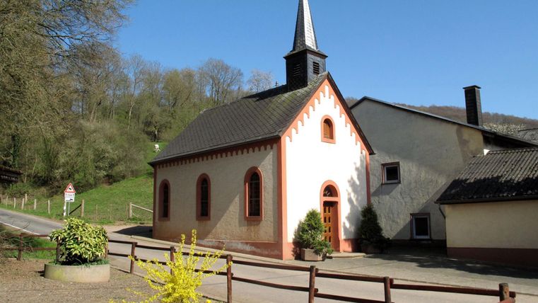 Une petite église avec un clocher pointu dans un environnement rural. Au premier plan, quelques fleurs et une rue bordée de bois sont visibles.
