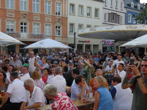 Eine belebte Platzsituation mit vielen Menschen, die an Tischen sitzen und miteinander feiern. Einige Gäste halten Getränke in der Hand und das Ambiente wirkt fröhlich und gesellig.