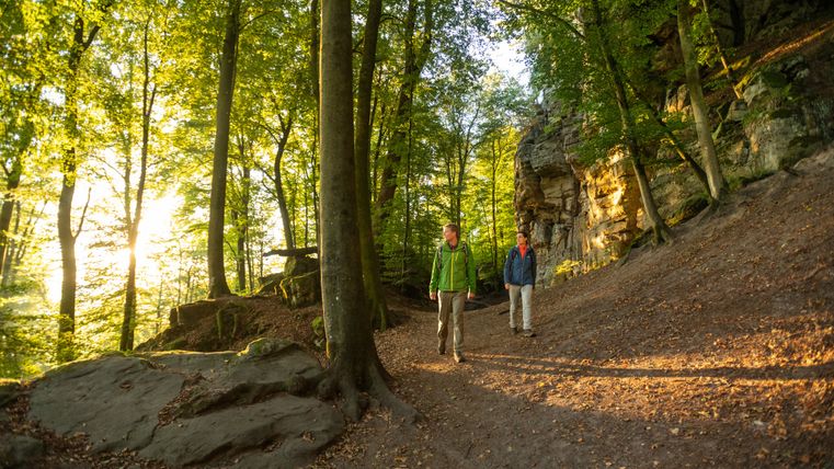 Zwei Personen wandern auf einem Waldweg nahe einer Felswand bei Sonnenuntergang.