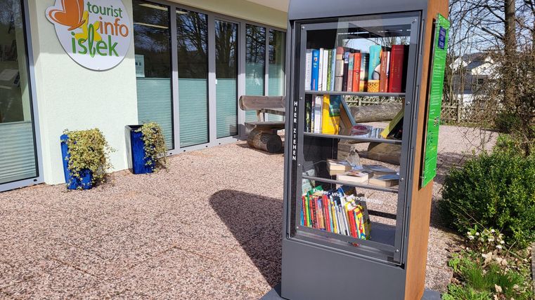 Une bibliothèque se trouve devant un centre d'information. Elle est remplie de différents livres et se situe dans un environnement verdoyant.