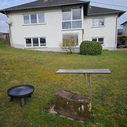 Een huis met grote ramen staat op de achtergrond. Op de voorgrond staan een tafel en een ronde grill op het gras.