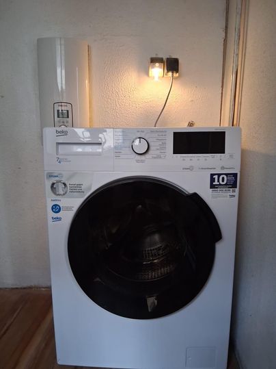 Une machine à laver moderne blanche est adossée à un mur. Elle a un grand hublot et un panneau de contrôle numérique.