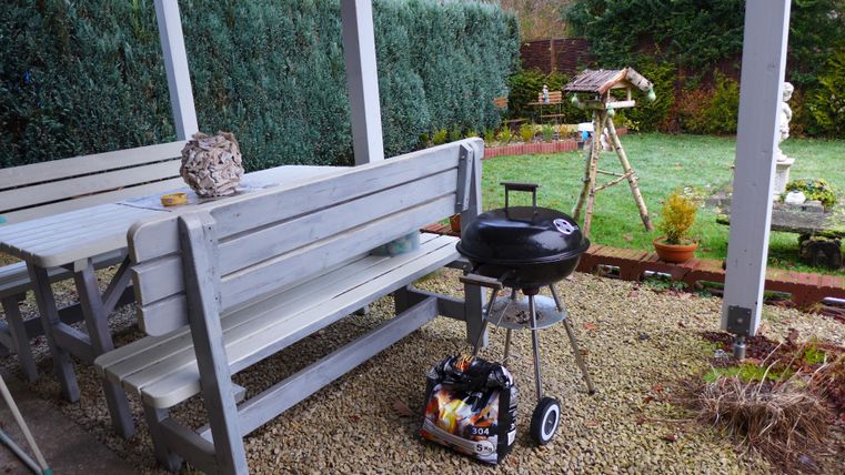 Eine gemütliche Terrasse mit einem Tisch und Bänken aus Holz. Darauf steht ein Grill und der Garten ist gut gepflegt.