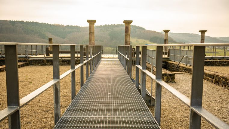 Ein Gehweg aus Metall führt durch eine archäologische Stätte mit mehreren Säulen. Im Hintergrund sind sanfte Hügel und ein bewölkter Himmel zu sehen.