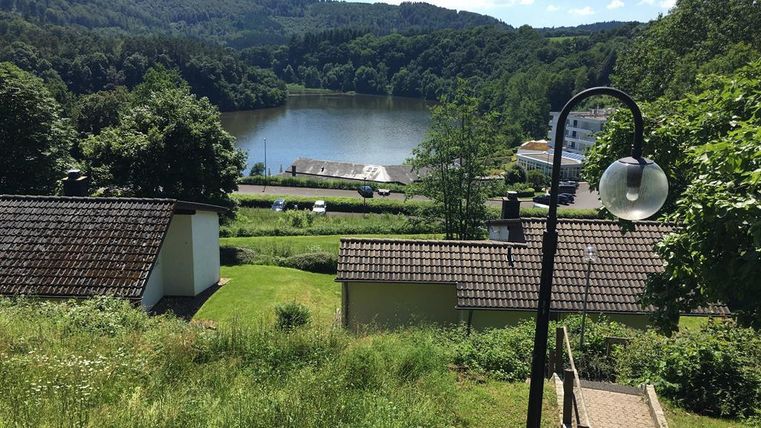 Un paysage pittoresque avec un lac calme et une végétation luxuriante. À l'arrière-plan, des collines et quelques bâtiments sont visibles.