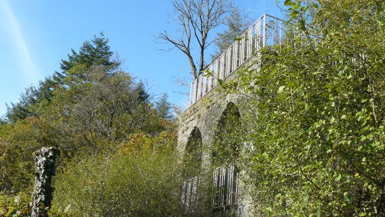 Mur de pierre avec balustrade dans un environnement boisé sous un ciel bleu.