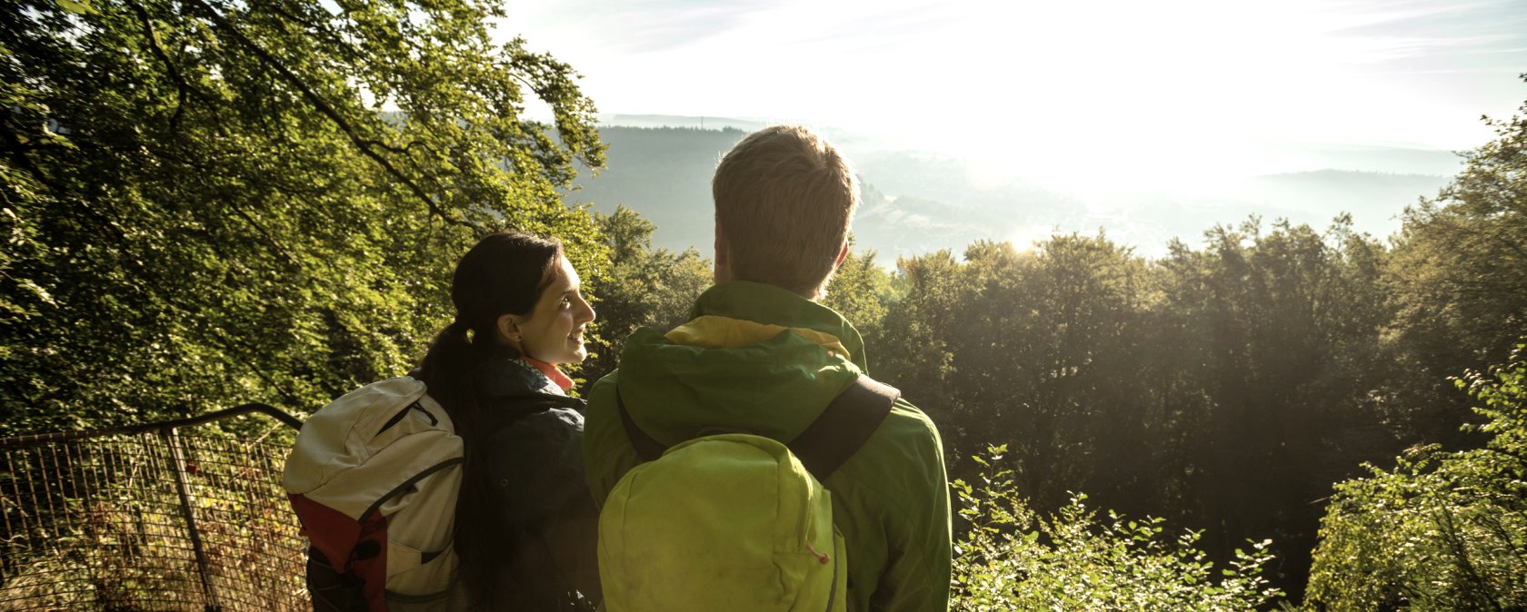 Zwei Wanderer mit Rucksäcken stehen an einem Geländer und blicken in die sonnige, bewaldete Landschaft., © Dominik Ketz