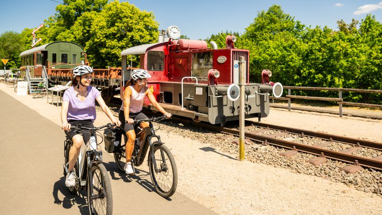 Zwei Radfahrerinnen fahren auf einem Radweg an einer alten Lokomotive vorbei.