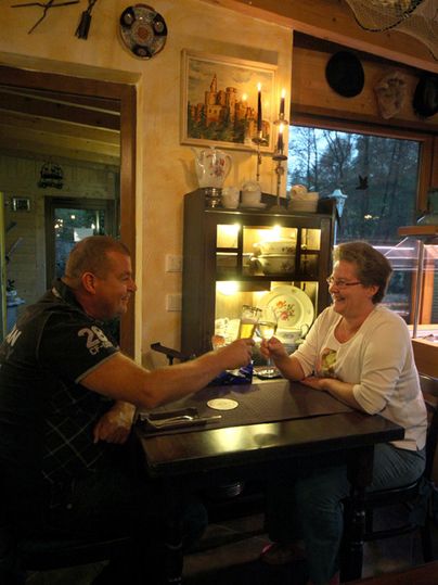 Un homme et une femme sont assis à une table dans une pièce confortable. Ils semblent discuter et rire, tandis qu'en arrière-plan, des objets décoratifs sont visibles.