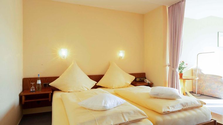 Ein gemütliches Zimmer mit zwei Betten, hellen Wänden und sanften Lichtquellen. Neben den Betten steht ein Nachttisch und es gibt einen Vorhang sowie eine kleine Pflanze im Hintergrund.