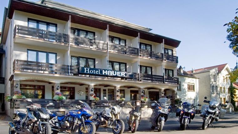 Un charmant hôtel nommé "Hôtel Hauer" avec plusieurs motos devant l'entrée. La façade de l'hôtel présente des balcons et de grandes fenêtres.