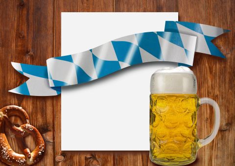 Ein Bierkrug mit goldenem Bier steht neben einer Brezel auf einem Holztisch. Im Hintergrund befindet sich ein leeres Blatt und eine dekorative blaue und weiße Schleife.