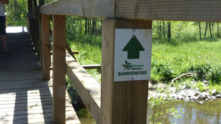 Ein Schild zur Barrierefreiheit, befestigt an einer gedeckten Holzbrücke über einem Fluss