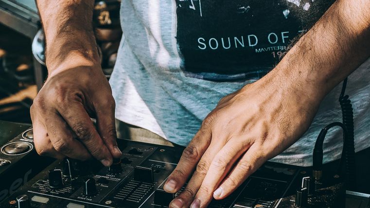 Ein DJ, der an einem Mischpult arbeitet. Die Hände sind auf den Reglern und Tasten des Geräts.