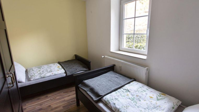 A simple room with two single beds and a window. The walls are painted light, and the floor is wooden.
