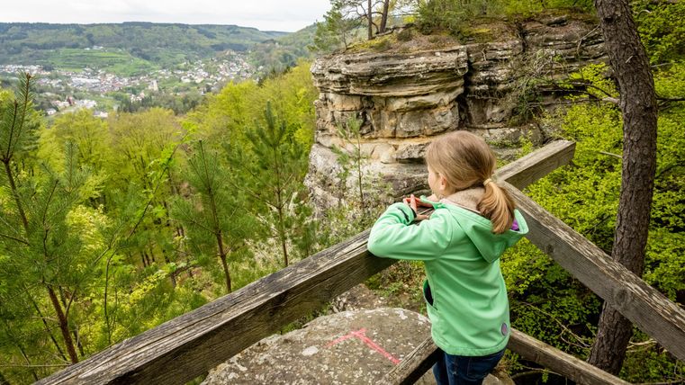 A girl stands at a railing and looks out over a green landscape with rocks and trees. In the background, hilly landscapes can be seen.