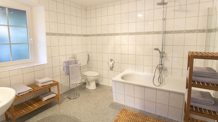 A modern bathroom with white tiles and a large window. There is a bathtub, a shower, and wooden shelves with towels.