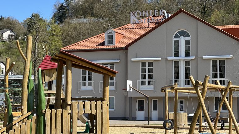 Ein Spielplatz mit Holzkonstruktionen steht vor einem zweigeschossigen Gebäude. Im Hintergrund ist eine Berglandschaft und ein blauer Himmel zu sehen.