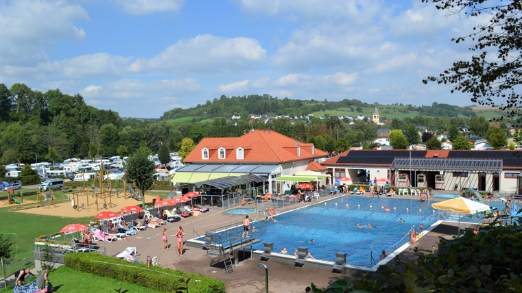 Ein großer Pool mit schwimmenden Gästen, umgeben von Liegestühlen und Sonnenschirmen. Im Hintergrund sind grüne Hügel und ein Campingplatz zu sehen.