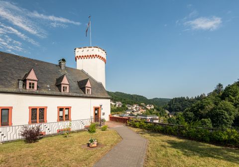 Ein historisches Gebäude mit einem Turm und einem hübschen Garten. Im Hintergrund sind grüne Hügel und ein kleiner Ort zu sehen.