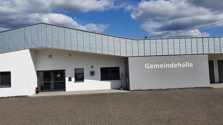 Ein modernes Gemeindezentrum mit einer klaren Fassade und großen Fenstern. Der Himmel ist blau mit vereinzelten Wolken.