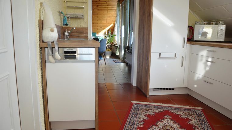 A modern kitchen with white cabinets and a carpet in the foreground. Windows and some furniture are visible in the background.