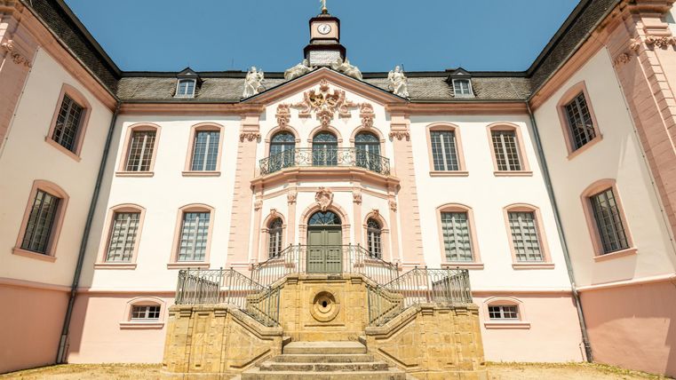 Ein historisches Gebäude mit einer eleganten Fassade in sanften Farben. Die große Treppe führt zu einem majestätischen Eingang mit Fenster und einem Uhrenturm darüber.